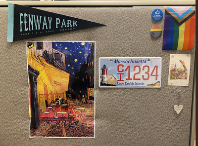 Items are pinned on a cubicle wall: a baseball pennant that says “Fenway Park,” a poster of  Vincent van Gogh’s painting “Cafe Terrace at Night,” a Cape & Islands Massachusetts license plate, a gay pride flag and pin, and various other mementoes.